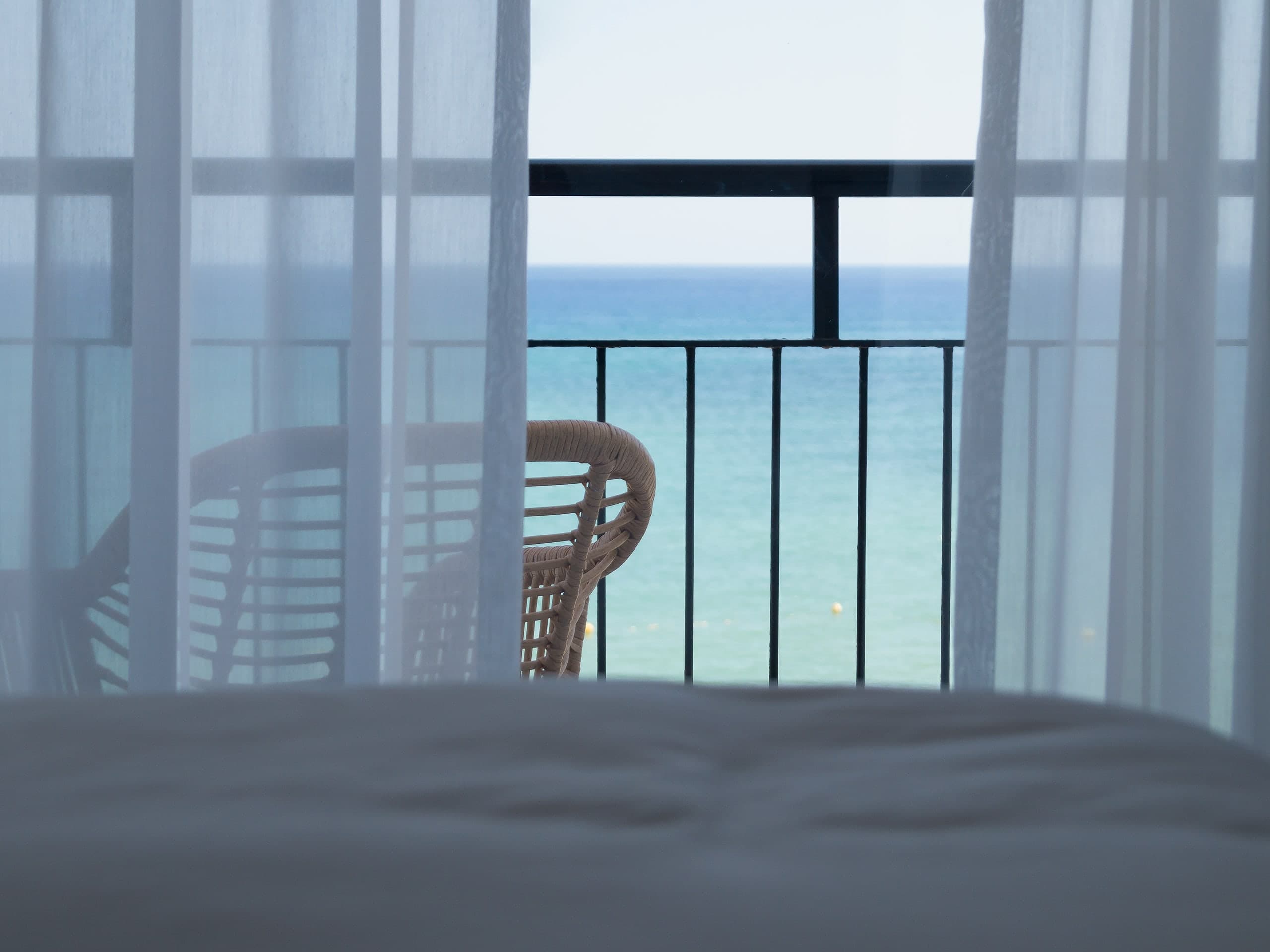 A hotel bedroom opening onto a sea-view terrace through sheer white curtains.